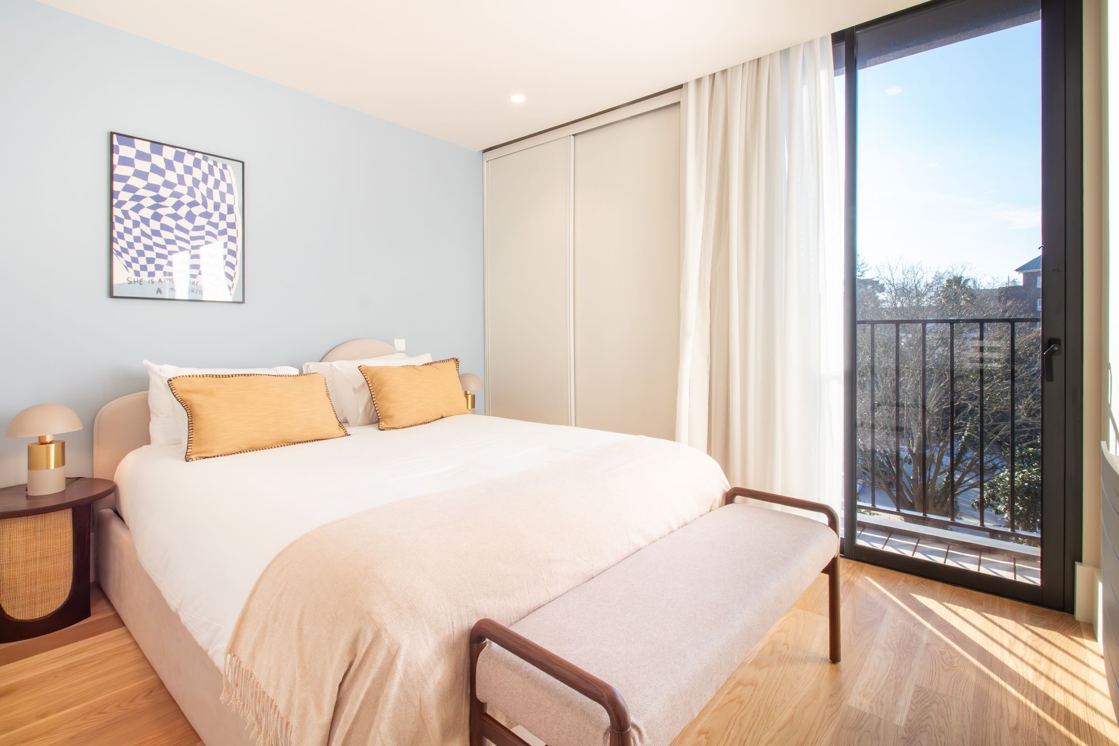 A bedroom with a large bed, a bench, a nightstand, a window with a view of trees, and a framed artwork on the wall.