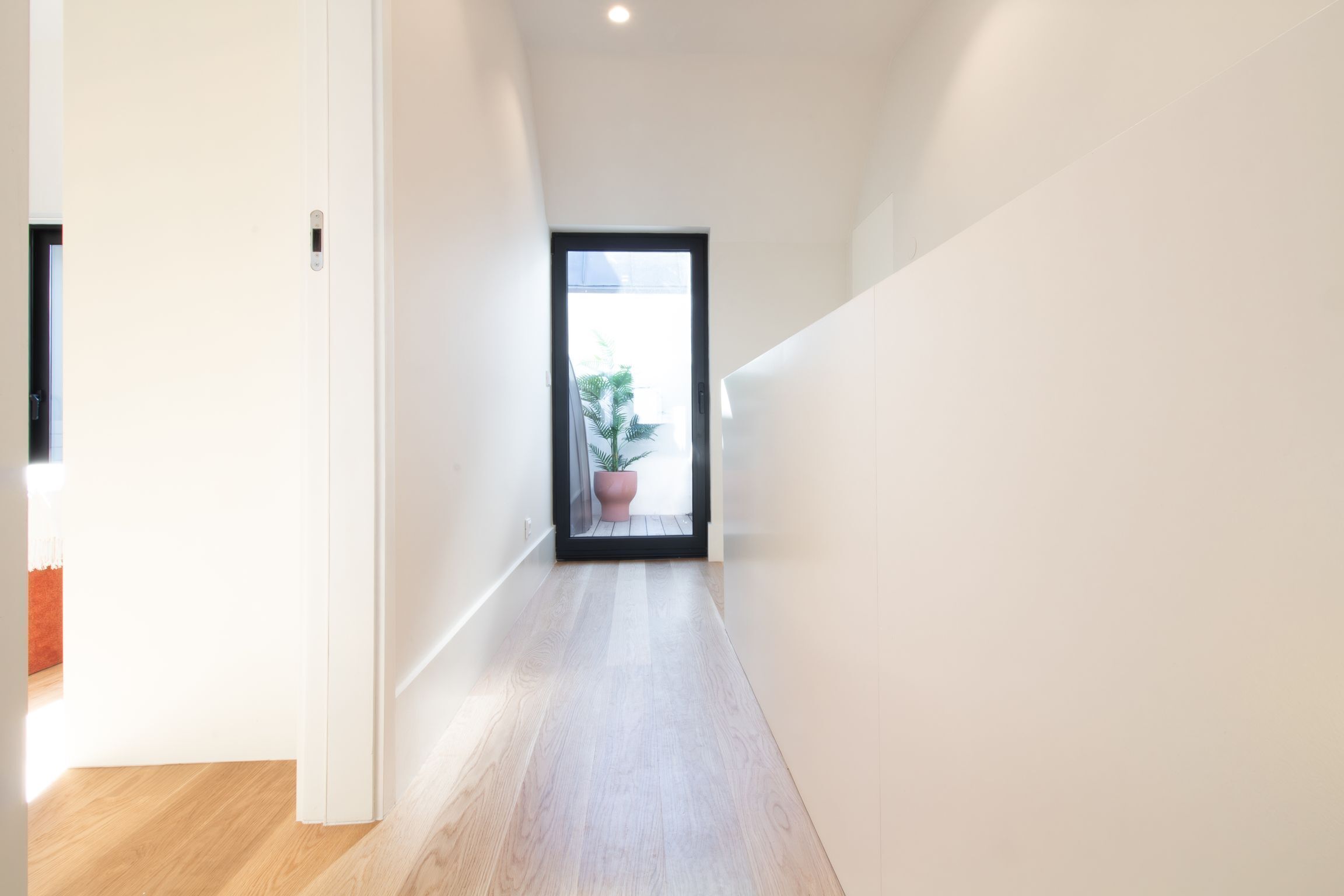 A narrow hallway with a wooden floor and white walls, leading to a door with a view of a potted plant outside.