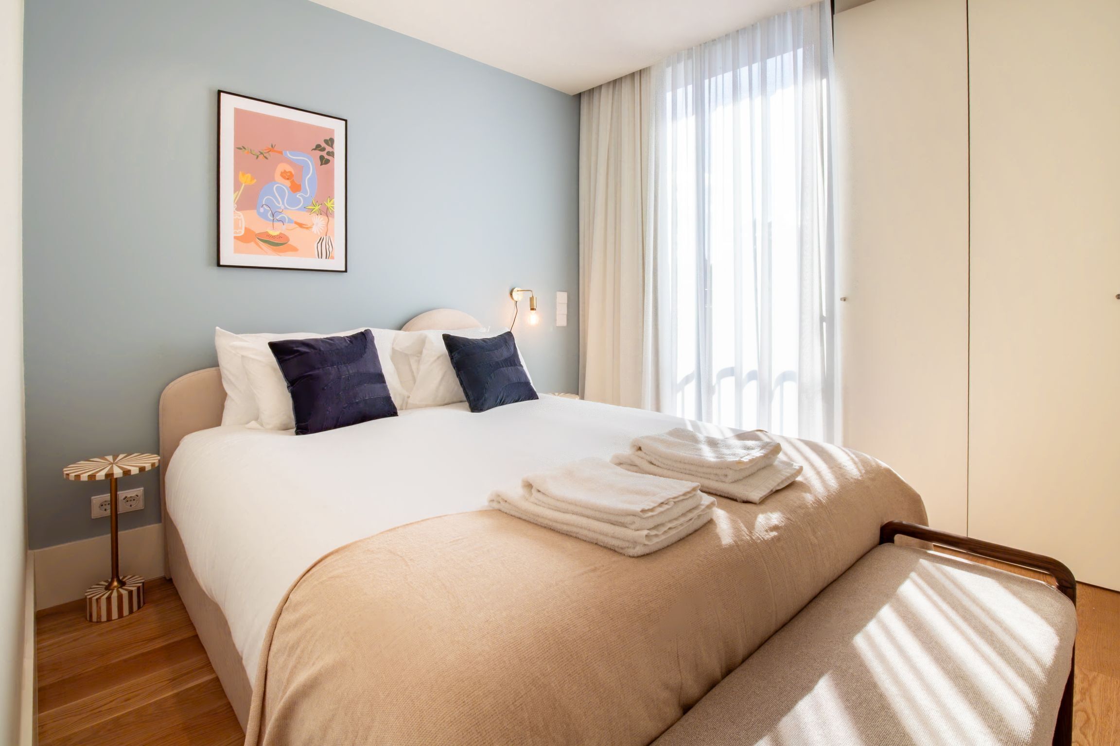 Un lit bien fait, avec des oreillers et une serviette pliée, une table de chevet avec une lampe et une œuvre d’art encadrée accrochée au mur.