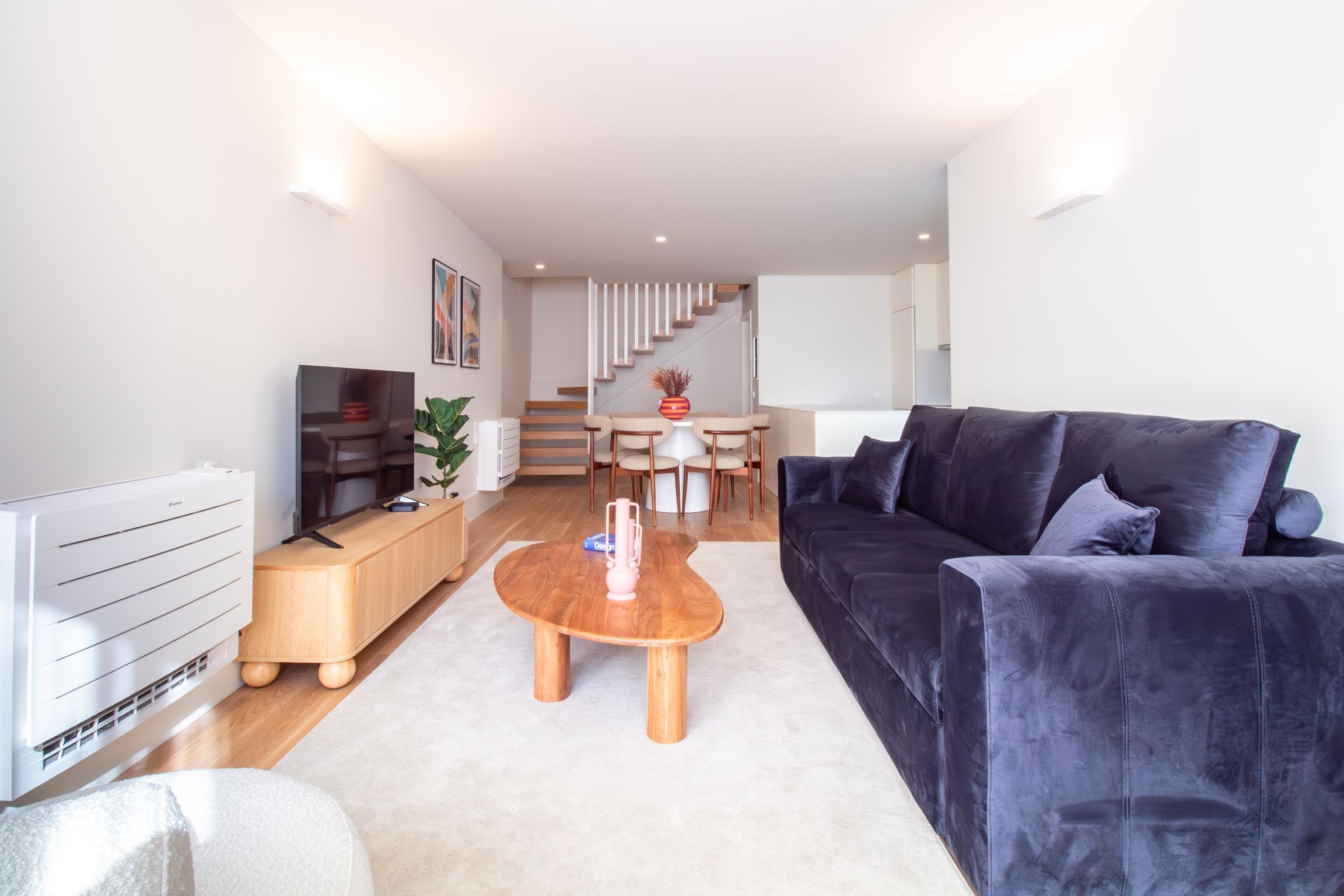 A modern living room with a dark blue sofa, a wooden coffee table, a white radiator, and a staircase leading to an upper floor.