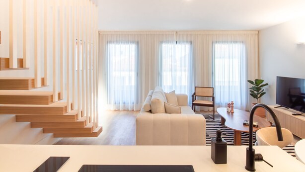 A modern living room with a sofa, a dining table, and a TV. There is a staircase on the left and a window with curtains. - Miguel Bombarda 259 by LovelyStay (Porto)