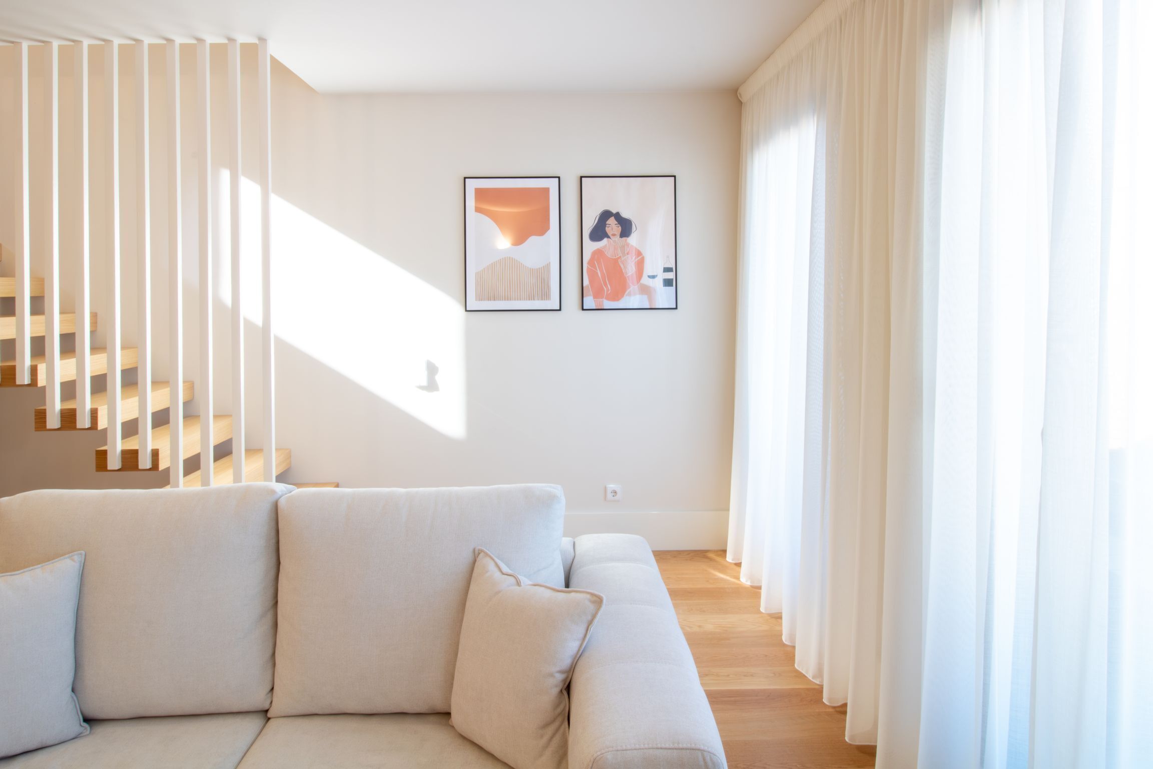 Uma sala de estar moderna com um sofá de cor clara, uma escada com degraus de madeira e duas obras de arte emolduradas na parede.