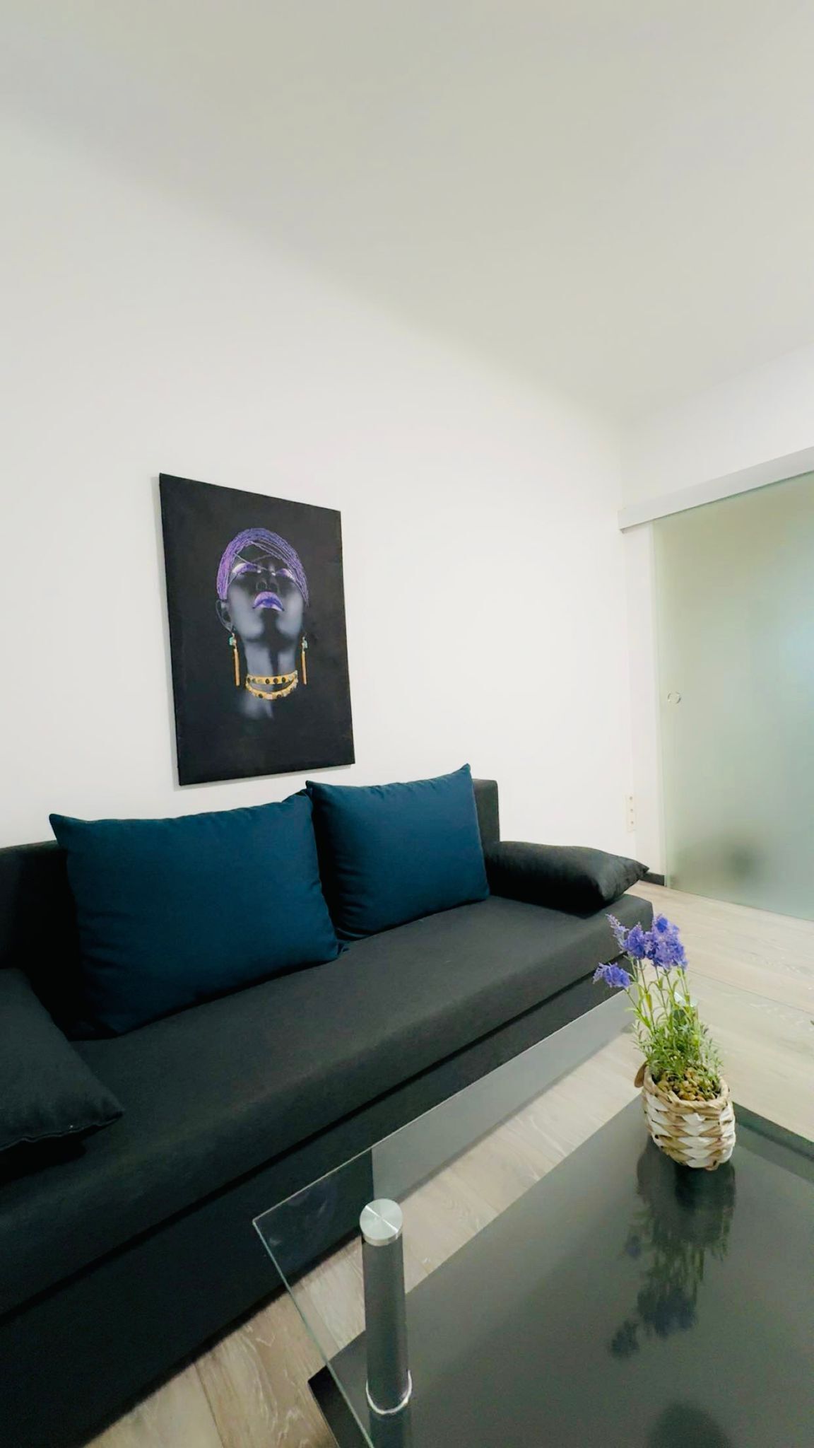 A modern living room with a black sofa, teal cushions, a glass coffee table, and a framed artwork on the wall.