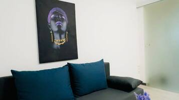 A modern living room with a black sofa, teal cushions, a glass coffee table, and a framed artwork on the wall.