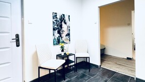 Une pièce avec un sol carrelé foncé, deux chaises blanches, une petite table sur laquelle est posée une plante en pot et une œuvre d’art accrochée au mur.