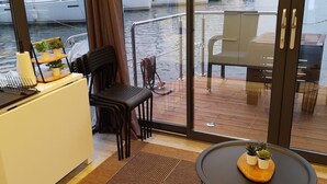 Ein kleines, kompaktes Zimmer mit Schreibtisch, Stuhl, Tisch und Blick auf das Wasser durch große Fenster.