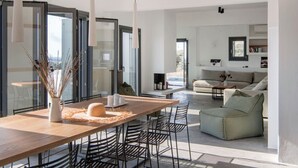 Un espacio habitable moderno con zona de comedor que incluye una mesa de madera y sillas de metal, un sofá de color claro y vistas a través de puertas de vidrio grandes.