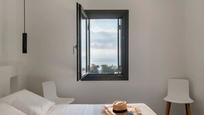 Un dormitorio con un ventanal grande, una cama, una mesita de noche con un sombrero y un libro, y una silla.