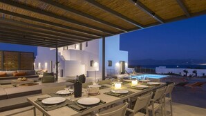 Un espacio de comedor al aire libre con piscina, paredes blancas y vistas a las montañas al atardecer.