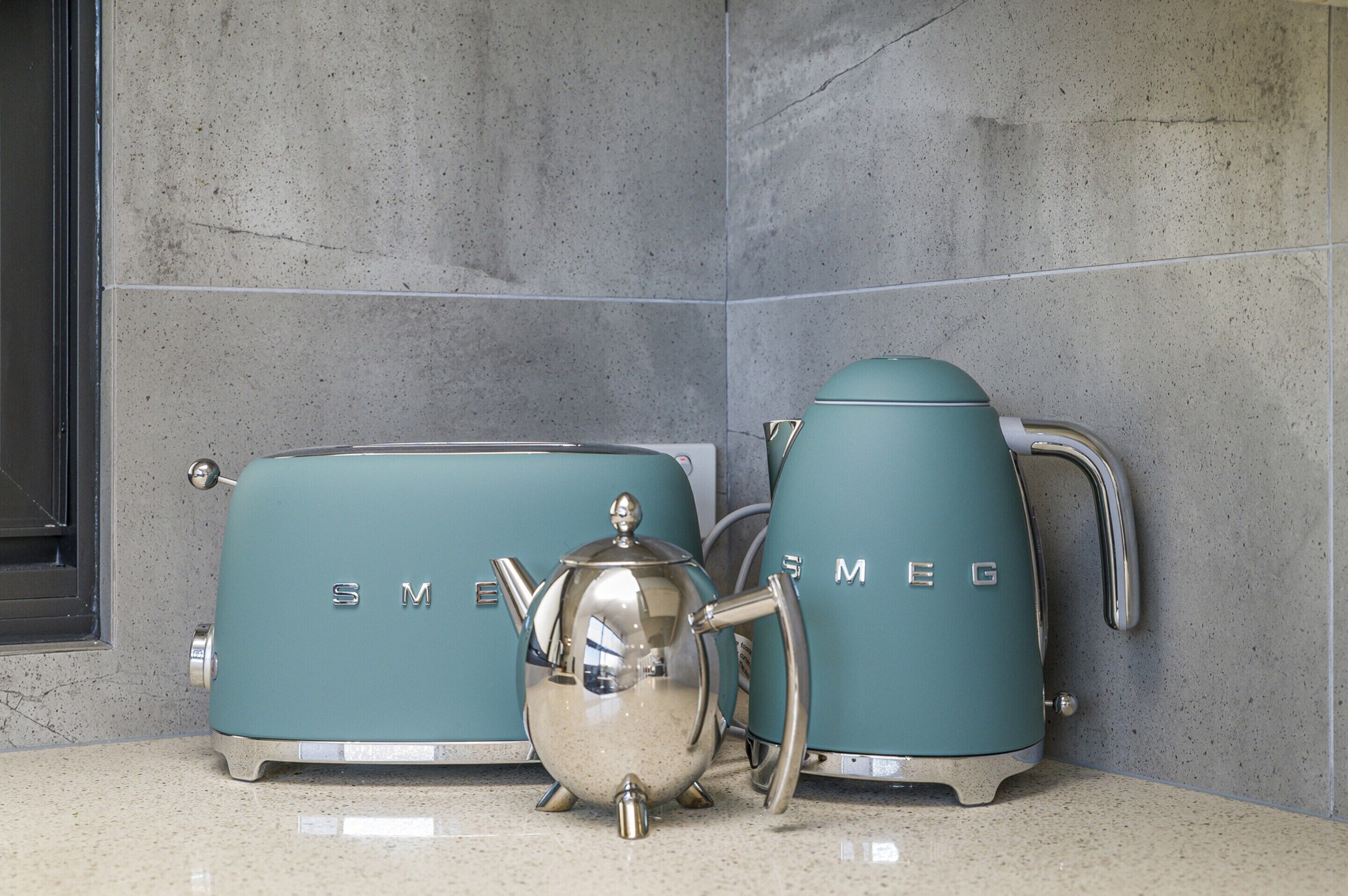 A kitchen with a stainless steel kettle and two teal appliances, likely a toaster and a coffee maker, against a tiled backsplash.