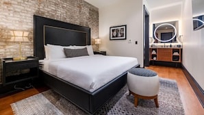 A modern bedroom with a brick wall, a large bed, bedside tables with lamps, a small stool, and a vanity with a mirror and lights.