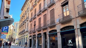Una calle flanqueada por edificios de varios pisos, algunos con balcones y escaparates, incluyendo una sucursal de CaixaBank.