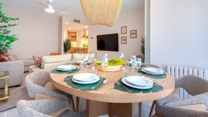 Un'area pranzo moderna con un tavolo rotondo in legno apparecchiato per sei persone, circondato da sedie grigie rivestite. Si notano un ventilatore a soffitto e un lampadario intrecciato sopra il tavolo.