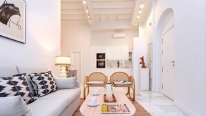Ein modernes Wohnzimmer mit einer Couch, einem Couchtisch und Sesseln. Im Hintergrund ist eine Küche mit weißen Schränken und Geräten zu sehen.