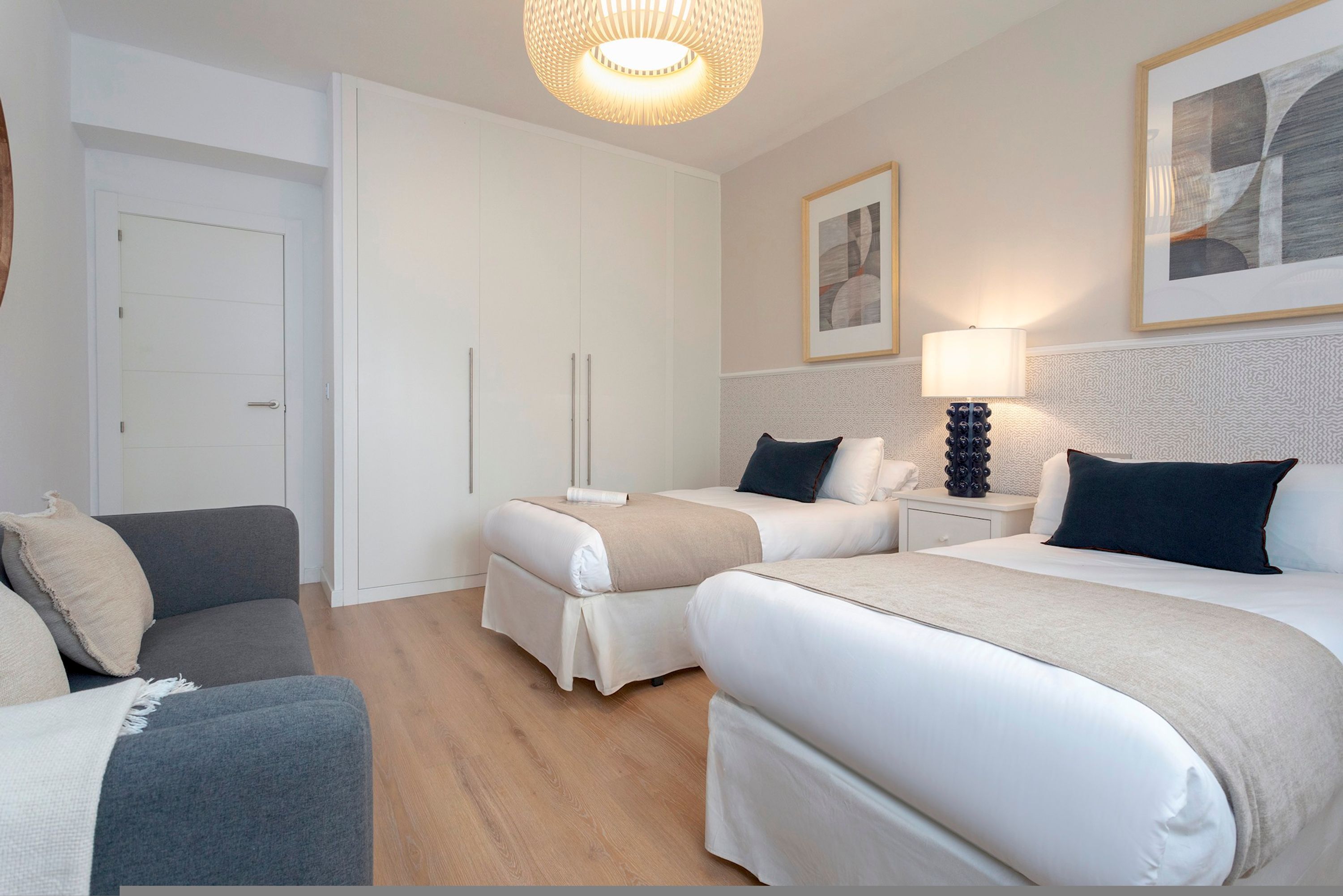 A hotel room with two beds, a sofa, a wardrobe, and a bedside table with a lamp.