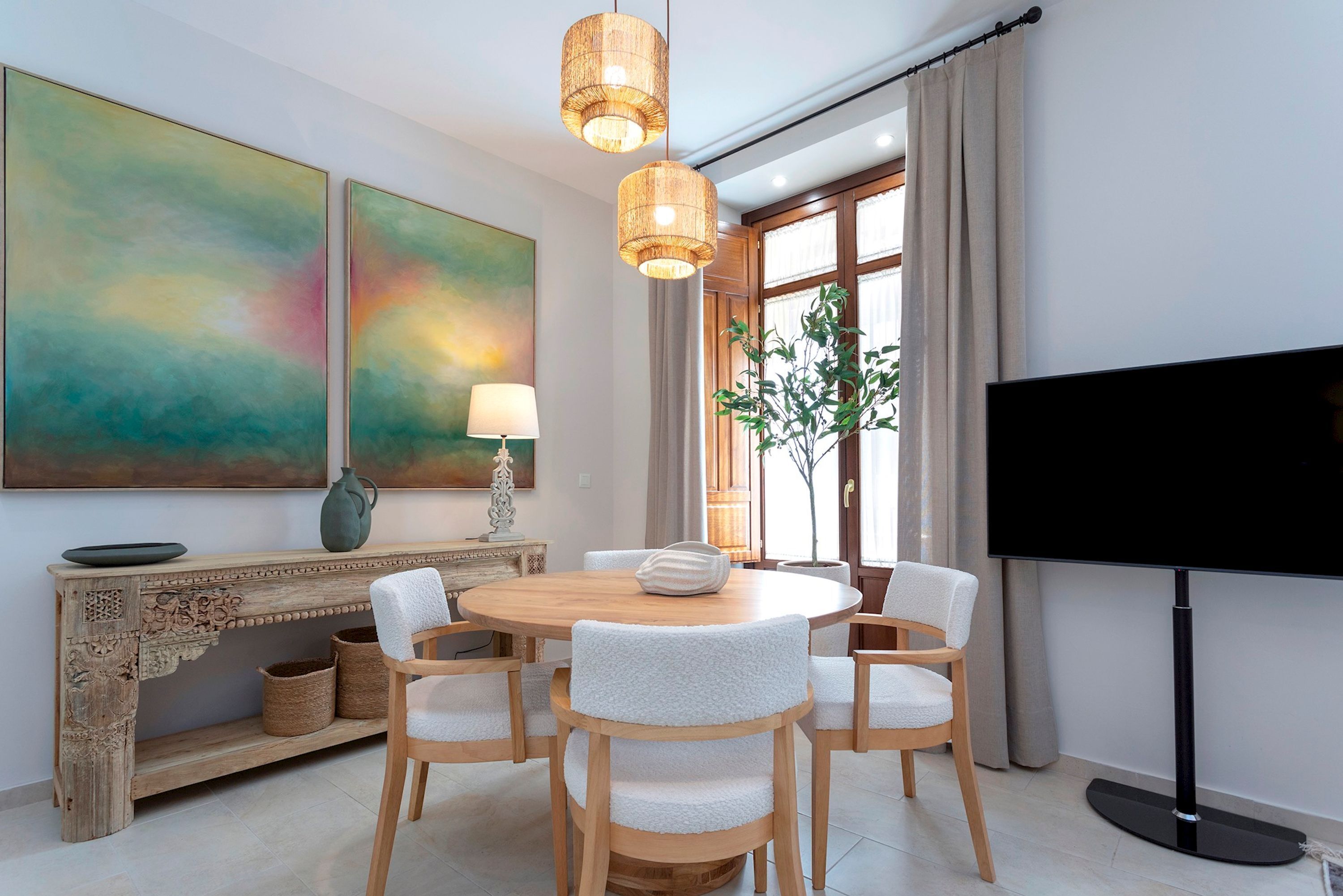A modern dining area with a round wooden table, six chairs, a large flat-screen TV, and two abstract paintings on the wall.