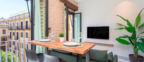 A balcony with a wooden table set for two, two chairs, a green sofa, a potted plant, and a TV mounted on the wall.