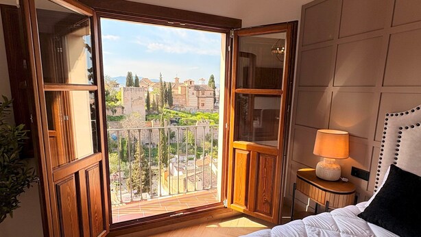 A bedroom with a large bed, a nightstand with a lamp, and a balcony with a view of buildings and greenery. - Lovely Loft With Views in Granada Mirador de Aixa (Granada)