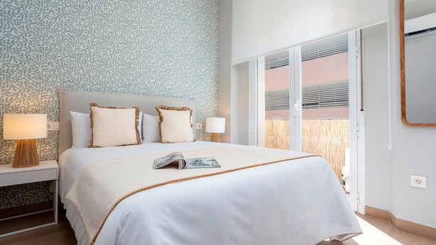 A neatly made bed with a white bedspread and two pillows, a bedside table with a lamp, and a window with blinds. - Cozy Central 2BD Apt in M Laga Trinidad Grund A (Málaga)