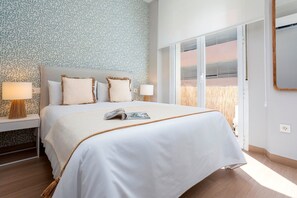 Uma cama bem arrumada com um cobreleito branco e duas almofadas, uma mesa de cabeceira com abajur e uma janela com persianas.