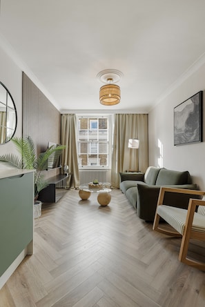 A modern living room with a herringbone wood floor, a green sofa, a wooden coffee table, and a large window with curtains. - Devonshire Terrace - 2 Bedroom Apartment (London)