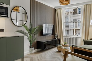 A modern living room with a flat-screen TV, a round mirror, a potted plant, and a window with curtains. - Devonshire Terrace - 2 Bedroom Apartment (London)
