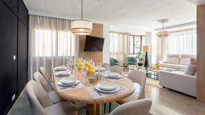 A modern dining area with a round table set for six, surrounded by comfortable chairs. A flat-screen TV is mounted on the wall, and there is a sofa and a coffee table in the background. - Luxury Apartment in City Center Puerta Real II (Granada)