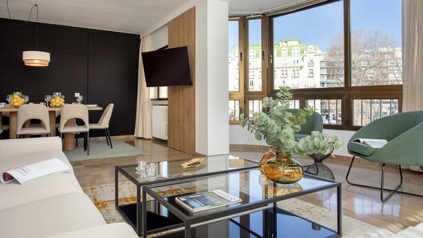 A modern living room with a glass coffee table, a sofa, a dining area with chairs, and a large window offering a city view. - Luxury Apartment in City Center Puerta Real II (Granada)