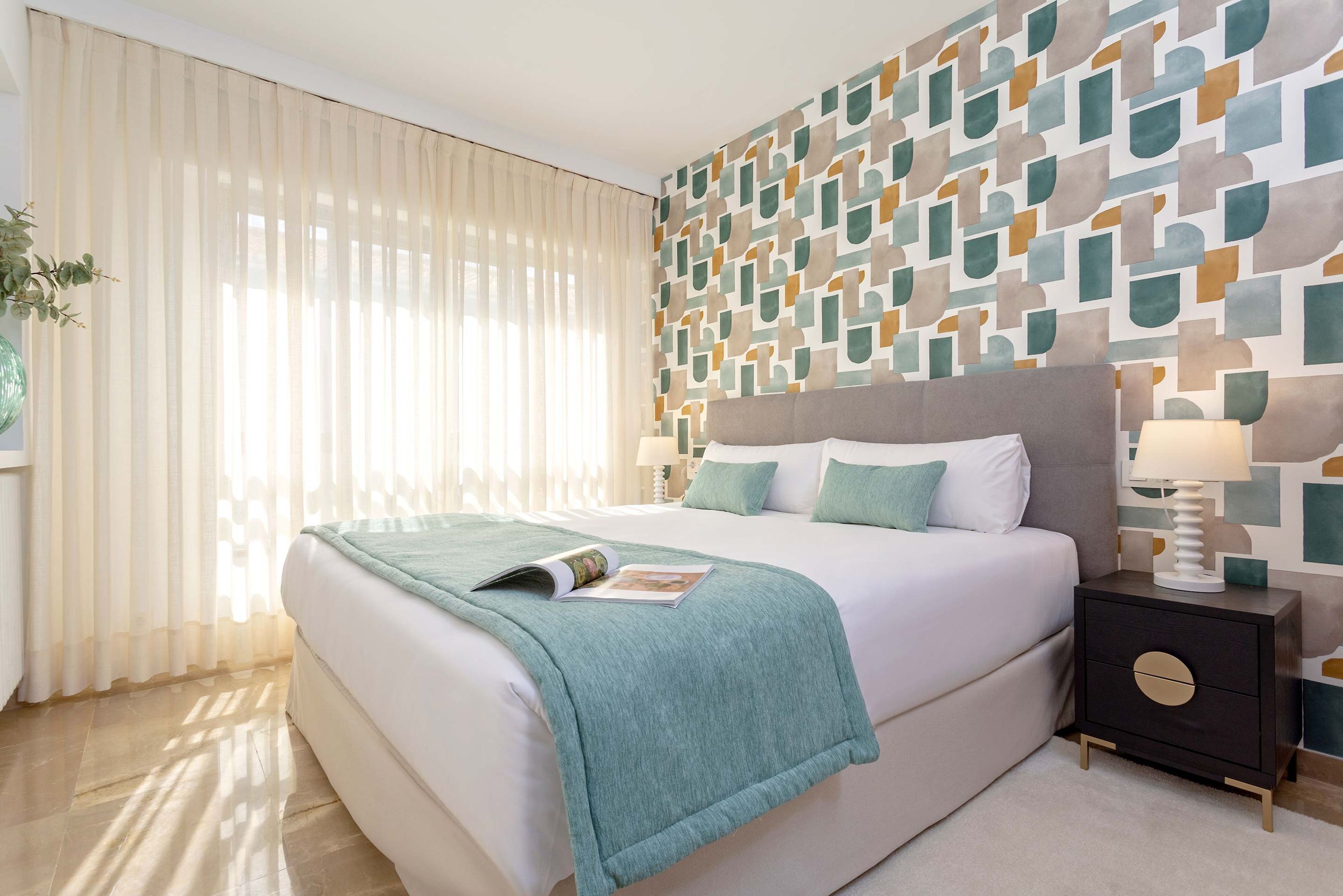 A modern bedroom with a large bed, a nightstand with a lamp, and a geometric-patterned wall.