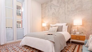 Ein Schlafzimmer mit einem Bett, zwei Nachttischen, einem kleinen Tischchen, einem gemusterten Boden und Blick auf ein Gebäude.
