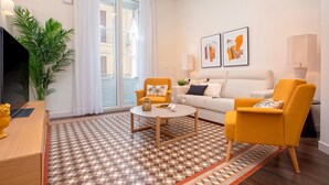 Ein modernes Wohnzimmer mit einer Couch, zwei gelben Sessel, einem Couchtisch und einem Fernseher. An der Wand hängen zwei gerahmte Kunstwerke, eine Topfpflanze und ein gemustertes Teppich.