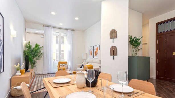 Ein Essbereich mit einem Holztisch, gedeckt für vier Personen, umgeben von Stühlen. Im Hintergrund befindet sich eine Couch und ein Couchtisch, sowie ein großes Fenster mit Vorhängen.