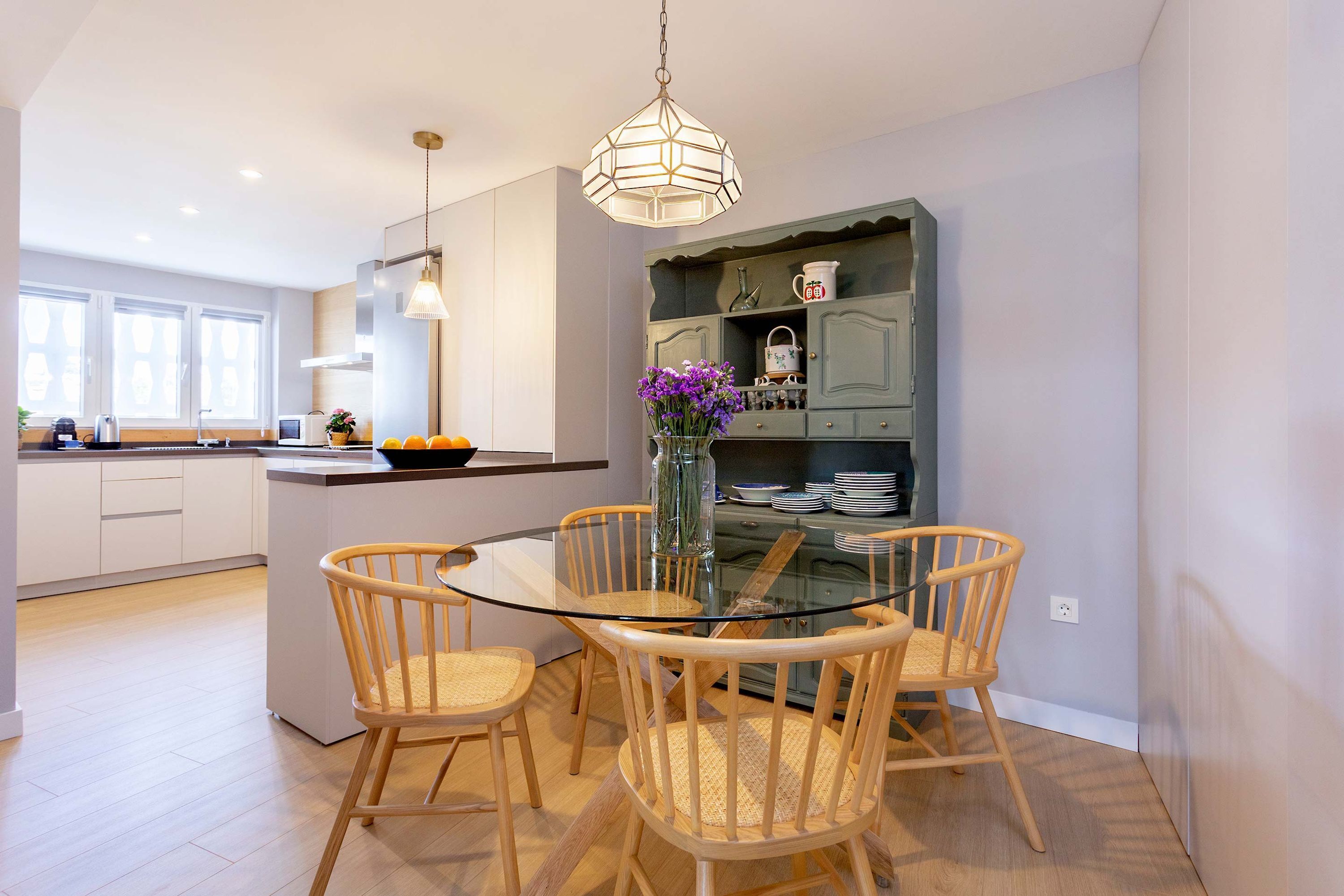 A modern kitchen with a glass dining table surrounded by wicker chairs, a hanging pendant light, and a built-in bookshelf with kitchenware.