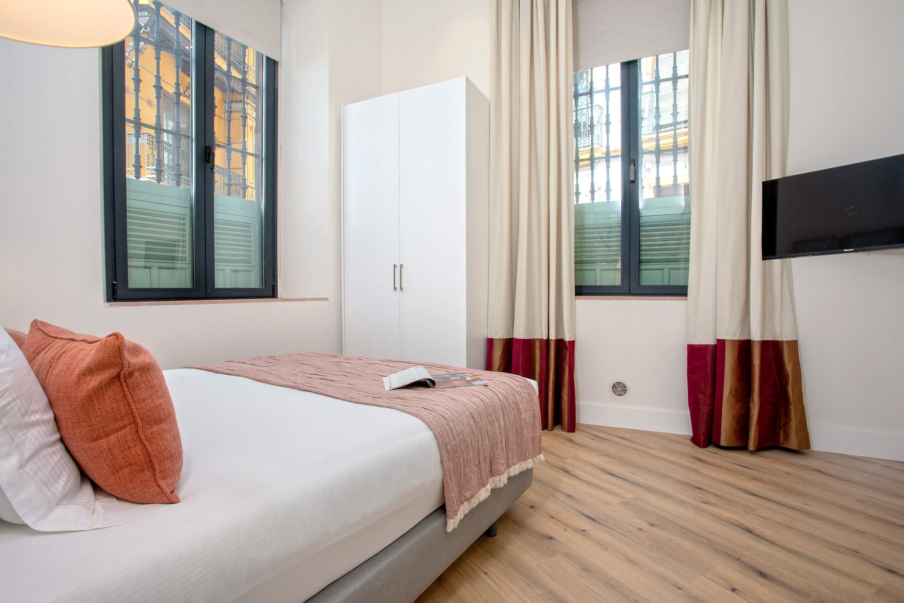 Un dormitorio con cama, armario, televisor y ventanas con cortinas.