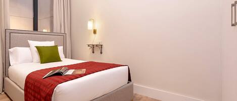 A neatly made bed with a red blanket, white pillows, and a green accent pillow. There is a bedside table with a lamp and a power outlet. The room has light wooden flooring and white walls.