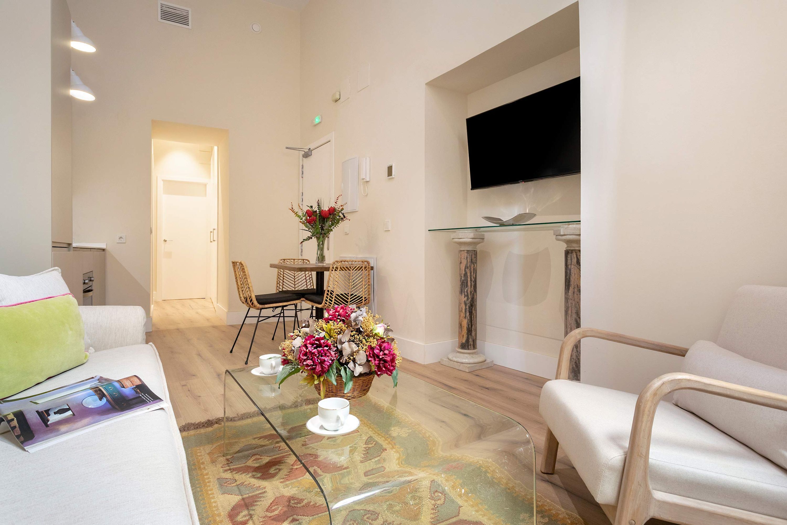 A modern living room with a glass coffee table, a sofa, a wicker chair, and a TV mounted on the wall.