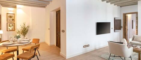 A modern living room with a dining area, featuring a round table, chairs, a flat-screen TV mounted on the wall, and a sofa.