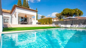 Uma piscina com superfície de água azul cristalina, rodeada por um edifício branco com telhado ladrilhado e uma área de varanda coberta com mobiliário de exterior.