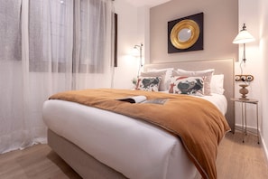 A neatly made bed with a brown blanket, white pillows, and a decorative pillow with a bird design. There is a round mirror on the wall above the bed and a small table with a lamp beside it. - Exclusive Apartment in Granada Horno Marina 9 (Granada)