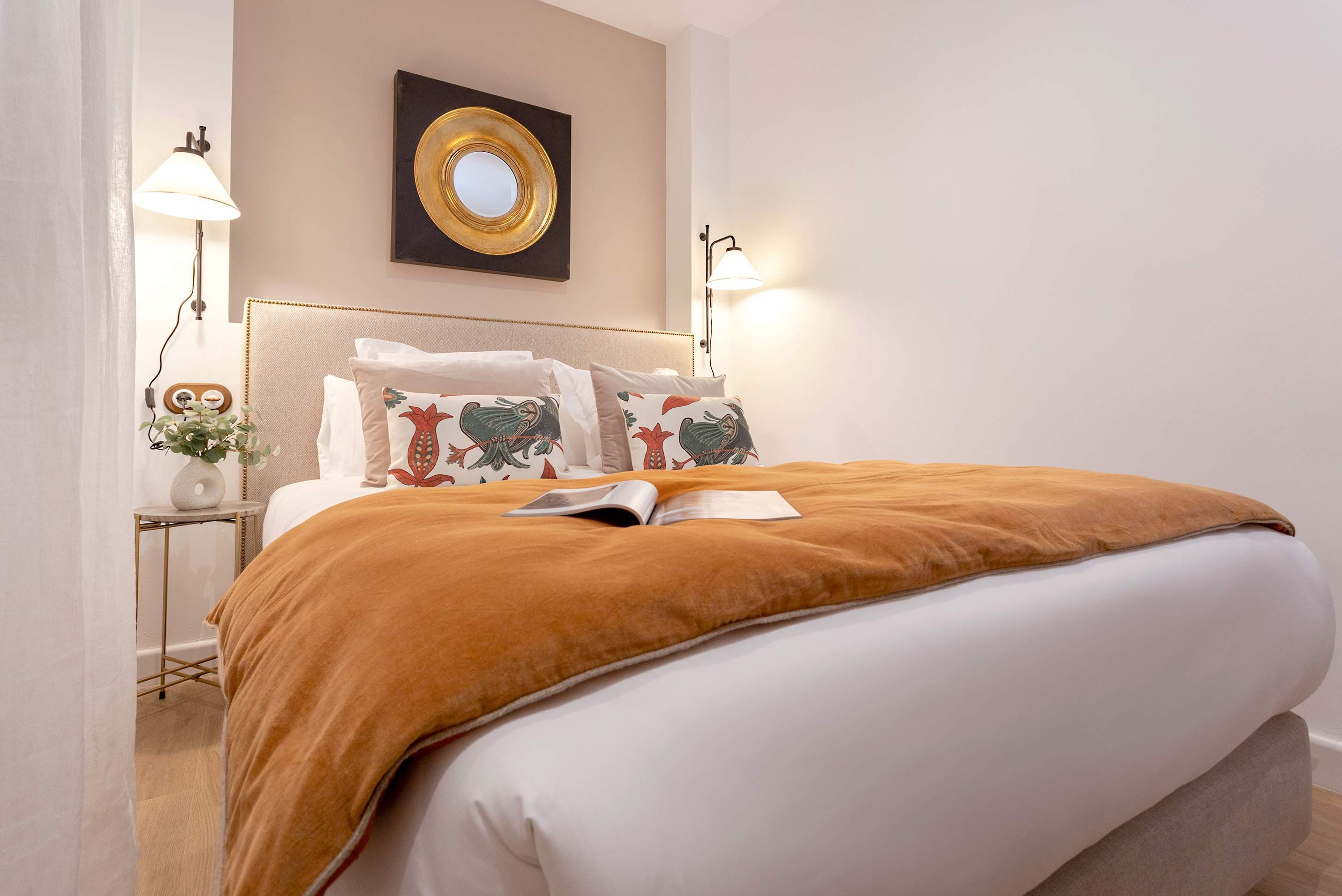 A neatly made bed with a brown blanket, two bedside lamps, a wall-mounted mirror, and a small table with a vase and a clock.