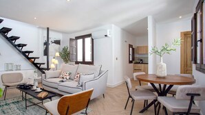 A modern living room with a grey sofa, a wooden dining table with chairs, a staircase, and a kitchen area visible through an open door. - Exclusive Apartment in Granada Horno Marina 11 (Granada)