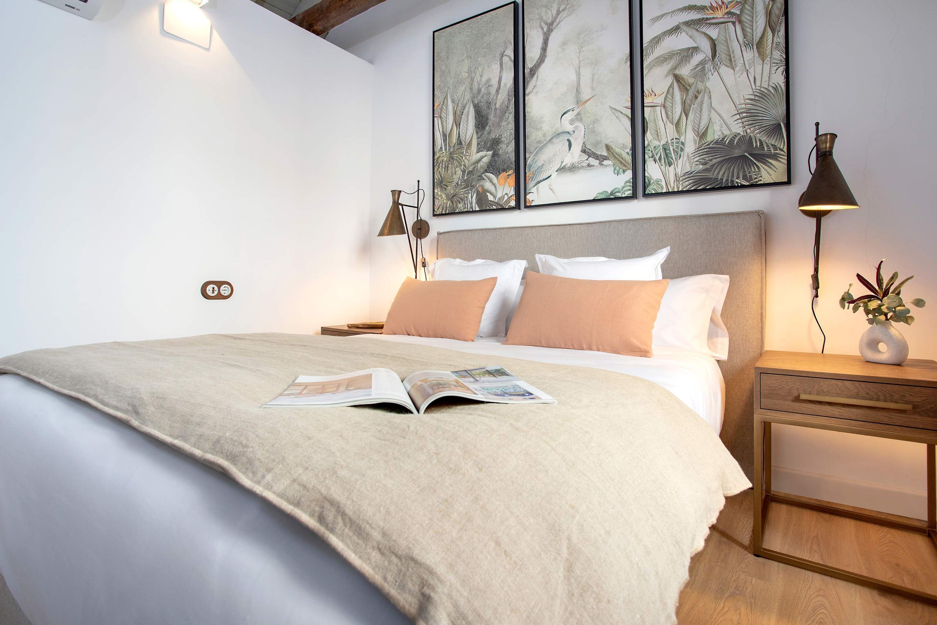 Una cama bien hecha con una revista, una mesita de noche con una lámpara y una pared decorada con cuadros.