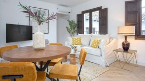 Un salón moderno con una mesa redonda de madera, sillas amarillas, un sofá blanco con cojines amarillos, un televisor y una planta.
