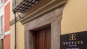 Entrada de un edificio con puerta de madera, un balcón con barandilla metálica y un letrero que dice "GENTEEL HOME HORNO MARINA".
