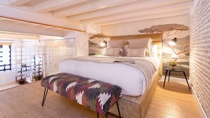 Une chambre avec un grand lit, un banc, une petite table avec une plante et un mur au motif de briques.