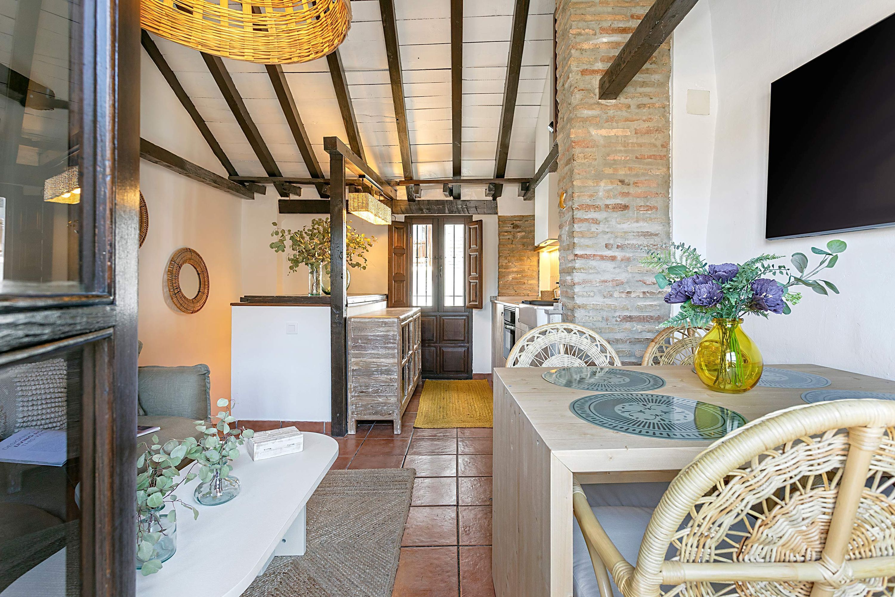 Ein rustikales Interieur mit freiliegenden Backsteinwänden, Holzbalken, einer Essecke mit Korbstühlen und einer Küche mit Ziegelrückwand.
