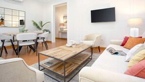 A modern living room with a wooden coffee table, white sofas, a dining area with chairs, and a flat-screen TV mounted on the wall.