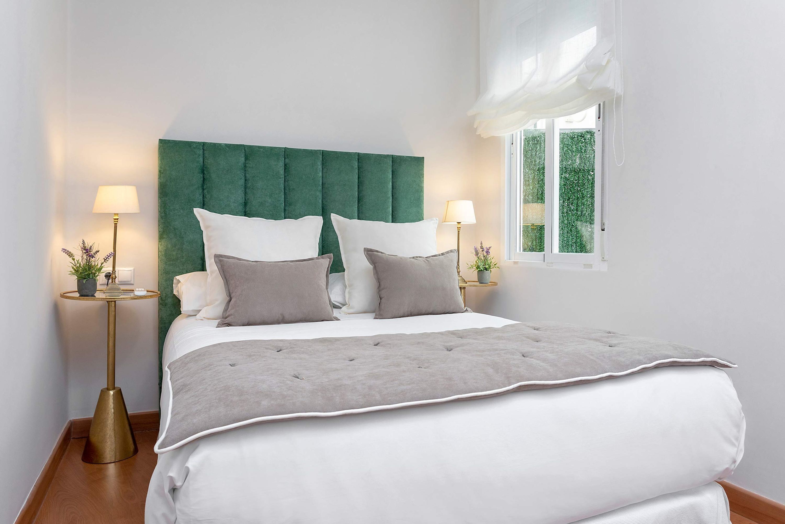 Un lit bien fait, doté d’une tête de lit recouverte d’un tissu vert, de draps blancs et gris, et de deux lampes de chevet sur des pieds dorés assortis.