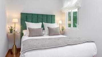 Un lit bien fait, doté d’une tête de lit recouverte d’un tissu vert, de draps blancs et gris, et de deux lampes de chevet sur des pieds dorés assortis.