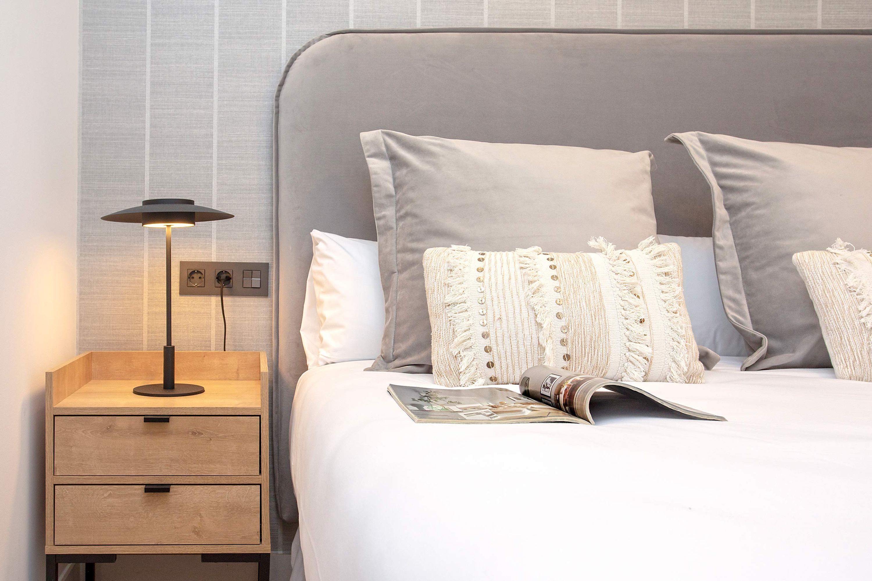 A neatly made bed with a magazine, a lamp, and a wooden nightstand with drawers.